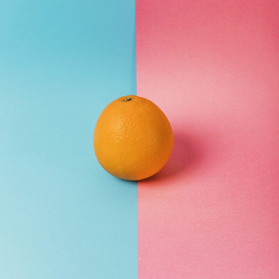 A single orange is positioned at the image's center, where a background split into two halves—light blue on the left and pink on the right—meets. Its brightness offers a striking color contrast against the pastel backdrop, enhancing web accessibility for all viewers