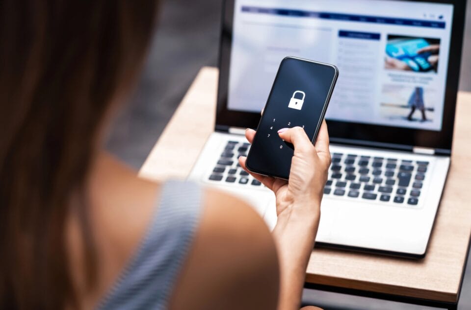 A person holds a smartphone, unlocking it with a passcode. In the background, a laptop displays a web page.