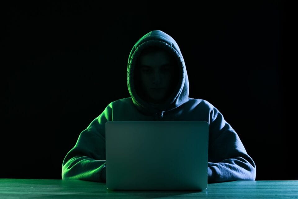 In a dimly lit room, a person in a hoodie sits before a laptop